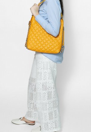 Vrouw in blauw shirt en witte kanten broek die een grote gele doorgestikte handtas over haar schouder draagt tegen een effen achtergrond.