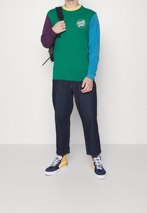 Camiseta de manga larga con cuerpo verde, mangas moradas y azules, cuello amarillo y logo en la parte delantera; combinada con jeans oscuros y zapatillas multicolores.