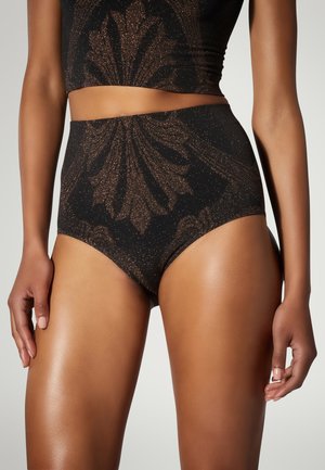 Sous-vêtement noir taille haute avec motif floral bronze scintillant porté par une personne debout, les bras relâchés le long du corps.