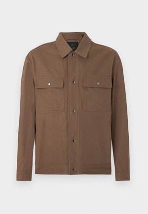 Brun knappet overskjorte med to klafflommer på brystet, lange ermer og klassisk krage på lys bakgrunn.