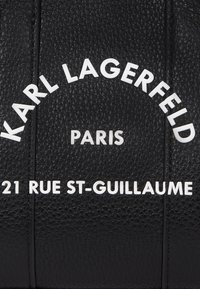Svart lærveske med preget tekstur, med hvit tekst som leser "KARL LAGERFELD PARIS 21 RUE ST-GUILLAUME" arrangert i et sirkulært mønster.