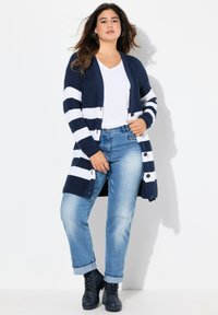 Pletený námořnický svetr s bílými vodorovnými pruhy, dlouhými rukávy, otevřeným předním dílem, kombinovaný s bílým topem s výstřihem do V a světle modrými džíny, doplněný černými botami.