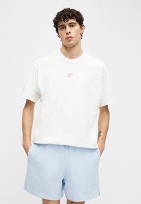 Νέος άνδρας με σγουρά μαλλιά, ντυμένος με λευκό μπλουζάκι Nike και ανοιχτό μπλε σορτς Nike, στέκεται με τα χέρια στις τσέπες μπροστά σε απλό φόντο.