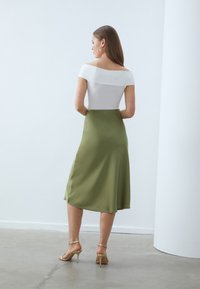 Haut blanc dénudé d'épaules associé à une jupe midi en satin vert, présentant une texture lisse et un design fluide, complété par des talons beiges à lanières.