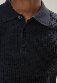 Navy Poloshirt mit strukturiertem Strickmuster, einer Drei-Knopf-Leiste und kontrastierendem Kragen. Weiches Gewebe mit subtilen geometrischen Details.
