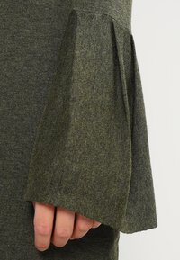 Olivgrön stickad tröja med utsvängda ärmar. Texturen är mjuk och har ett subtilt melerat mönster samt en avslappnad passform vid ärmarna.