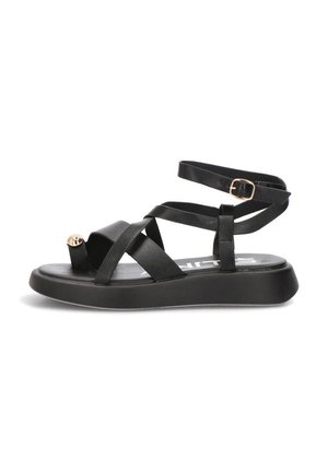Sandalia plana negra con tiras cruzadas, hebilla en el tobillo y una lazada en el dedo con punta dorada sobre una suela gruesa, vista de perfil.