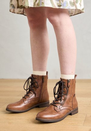 Beine mit cremefarbenen Socken und hellbraunen Leder-Schnürstiefeletten mit Seitentaschen, stehend auf Holzboden in einem geblümten Rock.