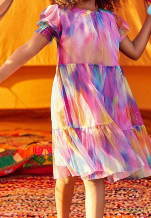 Enfant portant une robe colorée à motif galaxie pastel avec des manches bouffantes, debout sur un tapis tissé multicolore avec des coussins lumineux à proximité.
