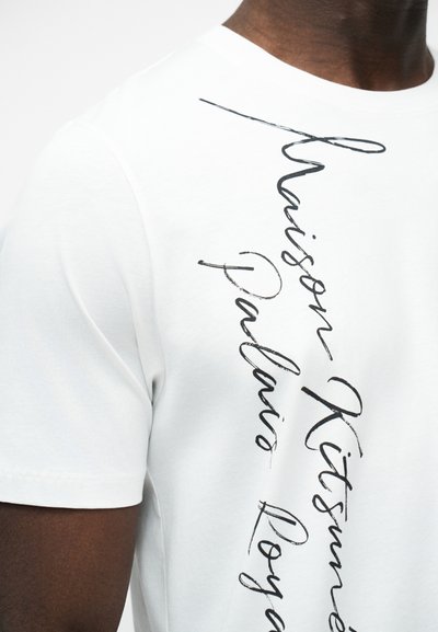 Λευκό μπλουζάκι με μαύρο κάθετο καλλιγραφικό κείμενο που γράφει "Thaison Kitsuné Palais Royal", φορεμένο από άτομο με σκουρόχρωμο δέρμα.