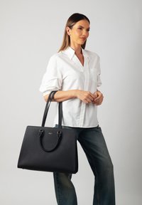 Black structured tote bag with a pebbled texture, gold hardware accents, and two long handles, paired with a white shirt and jeans.