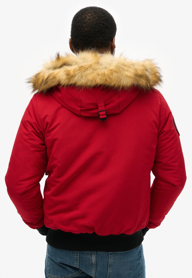 Veste doudoune rouge avec une capuche doublée en fourrure, des poignets côtelés et une taille élastique. Présente un petit logo à l'arrière et un design ajusté.