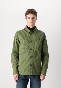 Olivově zelená bunda s límcem, zapínáním na knoflíky, dvěma náprsními kapsami a mírně strukturovanou tkaninou, nošená přes černý rolák.