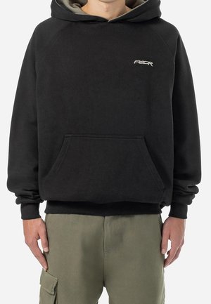 Hombre con sudadera negra con capucha y bolsillo canguro, con pequeño logo blanco en el pecho, combinado con pantalones cargo verde oliva.