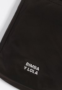 Czarna powierzchnia materiału z białym haftowanym napisem "BIMBA Y LOLA" w pobliżu dolnego rogu, ukazująca szczegóły ściegu i teksturę.