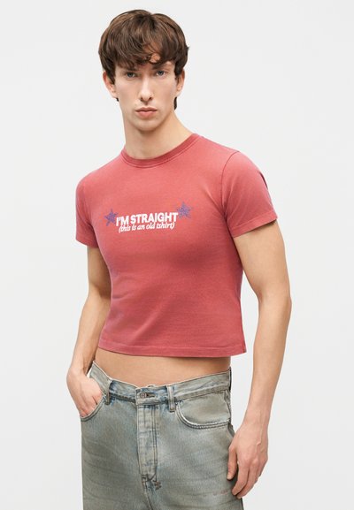 Mladý muž v krátkom červenom tričku s bielym textom „I'M STRAIGHT (toto je staré tričko)" a svetlom modrých džínsoch, stojaci na bielom pozadí.