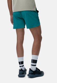 Teal træningsshorts, sorte sneakers med grå såler og hvide stribede sokker. En ensfarvet lys skjorte, set bagfra.