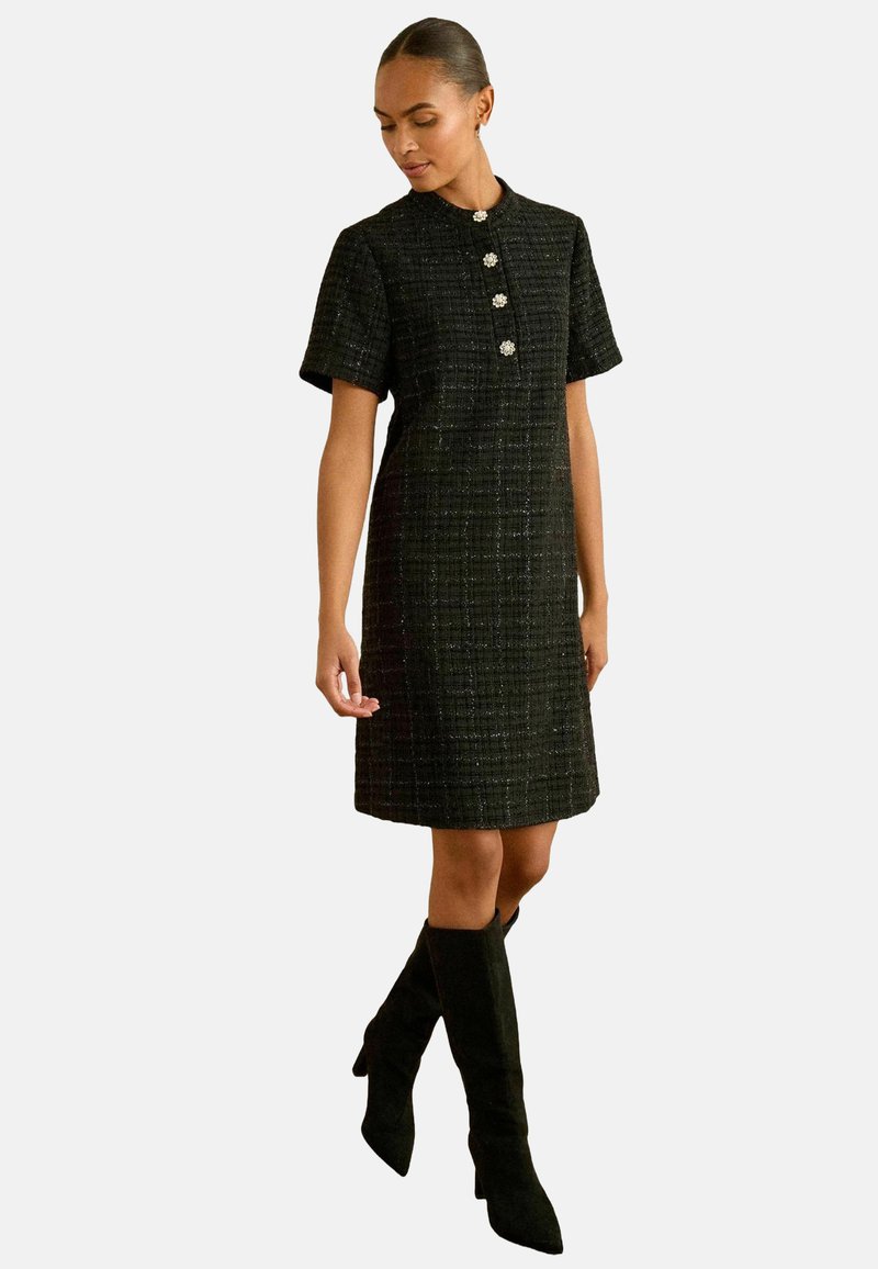 Musta tweed-mekko lyhyillä hihoilla, pyöreällä kaula-aukolla ja kolmella koristepainikkeella. Yhdistetty polvipituisille mustille saappaille, jotka näyttävät suoralta siluetilta.