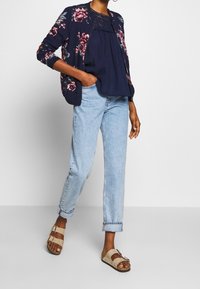 Blommönstrad marinblå kofta över en marinblå topp, tillsammans med ljusblå avslappnade jeans och beigea slidesandaler med två spännen.