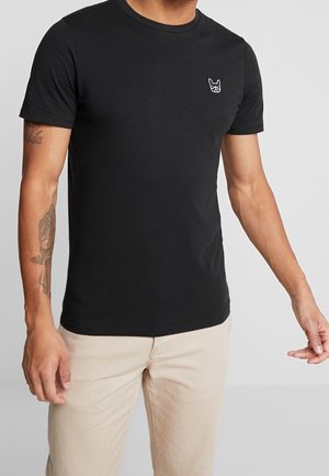 Mężczyzna w czarnej koszulce z krótkim rękawem z małym białym logo psa na piersi i beżowych spodniach, pokazujący tatuaże na lewym ramieniu.