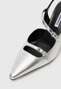 Sølvmetallisk høyhælte sko med spiss tå, ankelrem og utskjæringsdesign. Har en jevn tekstur med justerbart spenne. 
