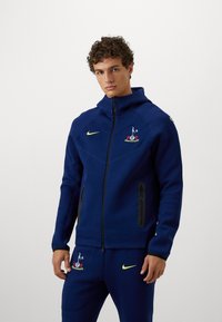 Mørkeblå atletisk jakke med højt krave, fuld lynlås og to sorte lynlåslommer. Har Nike-logo og broderet emblem på brystet.