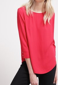 Blusa roja, ligera, de manga larga con escote en forma de barco, mangas de tres cuartos con detalles de botones y un dobladillo curvado.