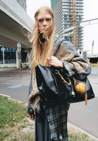 Jongere met lang blond haar, gekleed in gelaagde breigoed en geblokte kleding, houdt een grote zwarte leren tas vast met een gouden kettingaccessoire in een stedelijke omgeving.