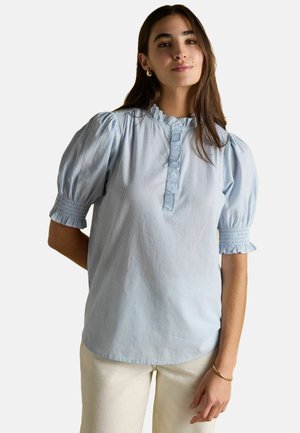 Woman with long brown hair wearing light blue short puff sleeve blouse with ruffled collar and white pants, standing against white background.