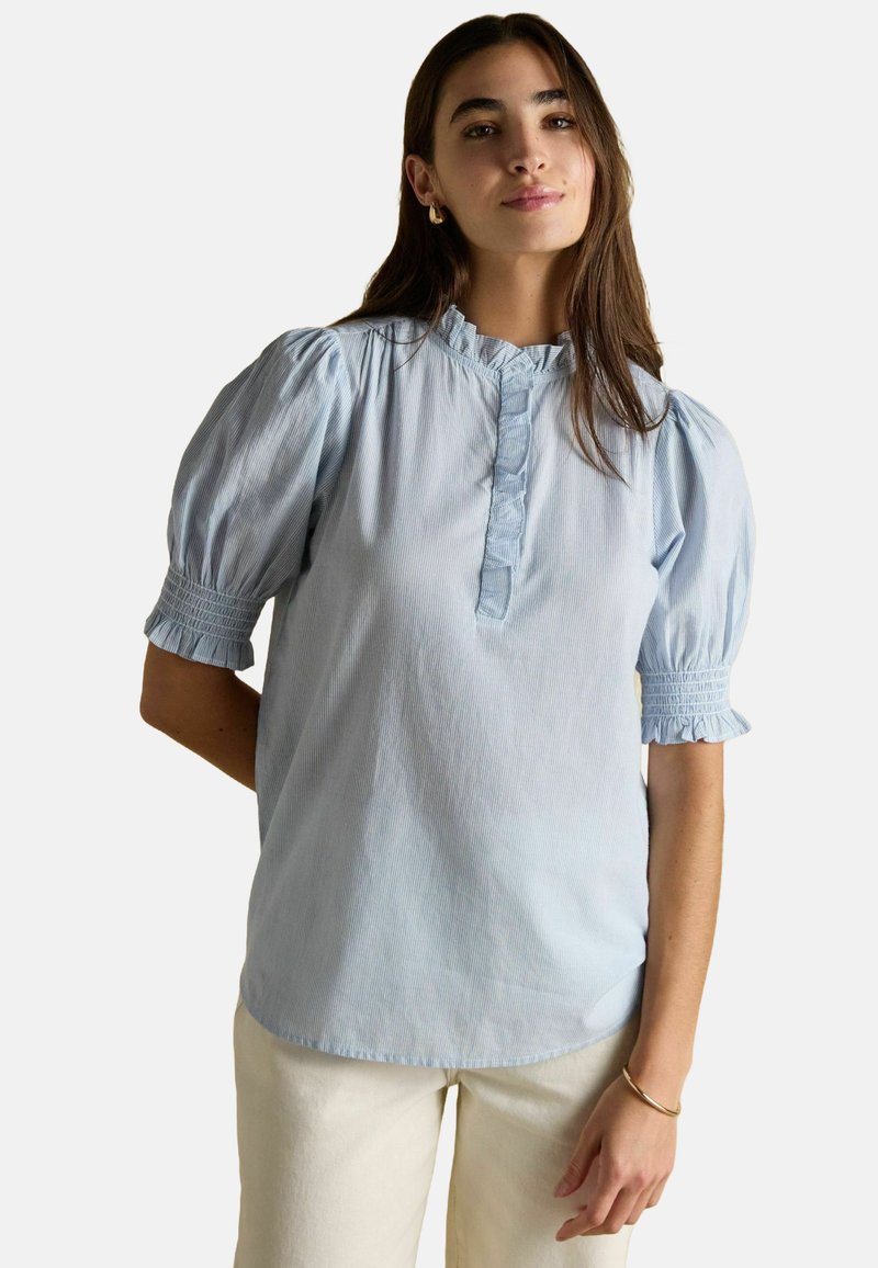 Frau mit langen braunen Haaren, die eine hellblaue Bluse mit kurzen Puffärmeln und Rüschensaum sowie weiße Hosen trägt, steht vor einem weißen Hintergrund.