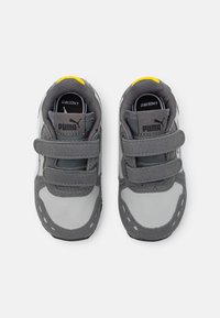 Grå och vita sneakers med kardborreband, med en texturerad yta, rundad tå och gult accent på hälen. PUMA-logotyp synlig.