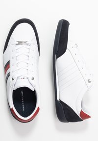 Vita låga sneakers med röda, svarta och grå ränder, svarta sulor och Tommy Hilfiger-logotyp på tungan och innersulan.