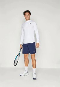 Biela športová košeľa s dlhým rukávom, úzkeho strihu a golierom, v kombinácii s námorníckymi šortkami. Model drží tenisovú raketu a má na sebe biele tenisky.