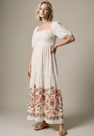Frau mit kurzen blonden Haaren, die ein weißes, gerafftes Kleid mit Puffärmeln und bunter floraler Stickerei am Saum trägt, steht mit der Hand in der Hüfte.
