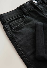 Gros plan sur un jean en denim noir plié montrant la poche avant, la boucle de ceinture, et le détail du bouton sur fond clair.