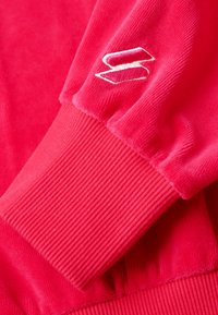 Gros plan d'une manche de sweat-shirt rouge avec un poignet côtelé et un logo stylisé "S" blanc brodé près du poignet.