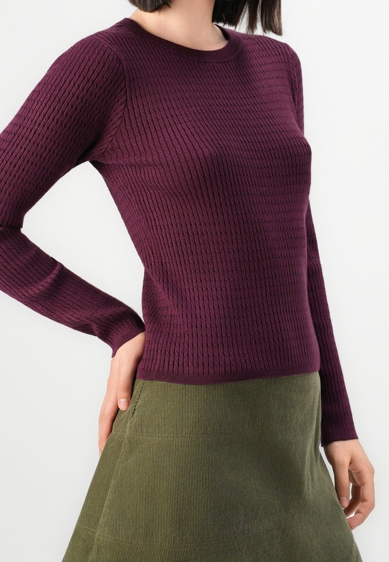 Femme portant un pull violet côtelé à manches longues et une jupe en velours côtelé vert olive, debout avec une main sur la hanche.