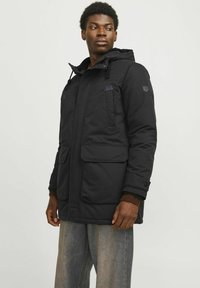 Schwarze gepolsterte Parkajacke mit Kapuze, ausgestattet mit mehreren Vorderaschen, einem Reißverschluss und gerippten Bündchen. Getragen über hellblauen Jeans.