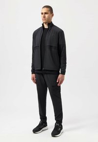 Black athletic tracksuit featuring a zip-up jacket with mesh panels and elastic cuffs, paired with tapered pants and black sneakers.