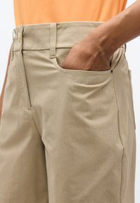 Χέρι στην εμπρόσθια τσέπη μπεζ παντελονιού που φοριέται με πορτοκαλί πουκάμισο, δείχνοντας την υφή του υφάσματος και τη λεπτομέρεια της τσέπης.