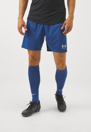 Mężczyzna ubrany w niebieskie spodenki Under Armour, niebieskie skarpetki do kolan Nike, czarne korki Nike i czarną koszulkę, stoi na jednolitym tle.