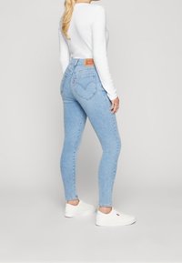 Ljusblå skinny jeans med hög midja, med en läderlogotyp på baksidan och subtila sömnadsdetaljer. Bärs med en vit långärmad skjorta.
