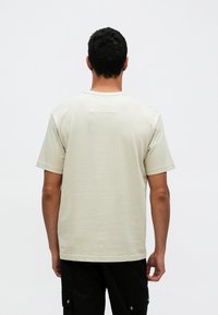 Šviesiai rudos spalvos trumpomis rankovėmis marškinėliai iš minkštos medžiagos, su apvalia iškirpte ir mažu kilpiniu ženkleliu nugarinėje dalyje, dėvimi su juodomis krovininėmis kelnėmis.