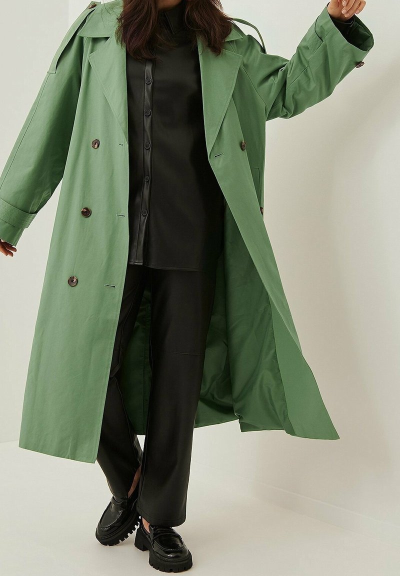 Kobieta w długim zielonym trenczu, nosząca czarną koszulę zapinaną na guziki, czarne spodnie i masywne czarne buty, stojąca na tle jednolitego białego tła.