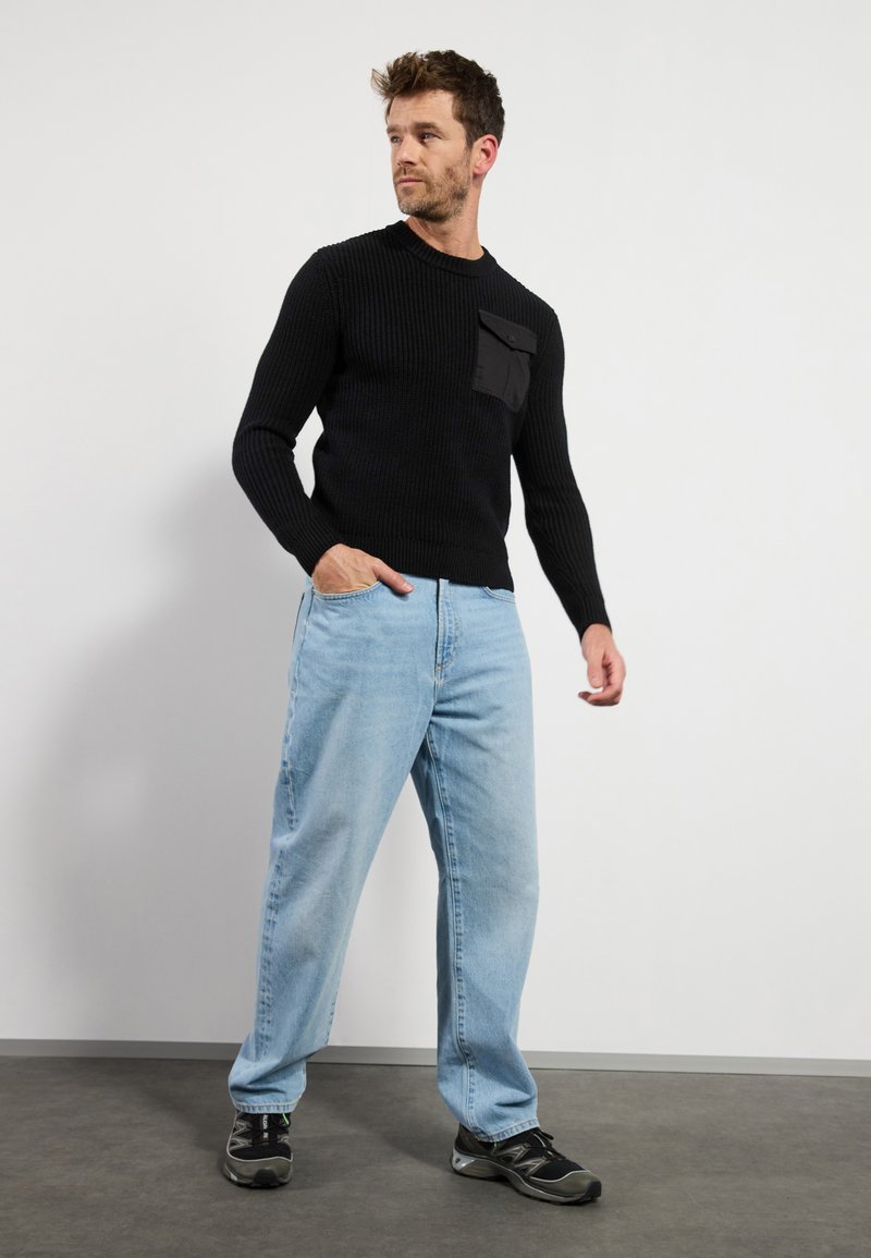 Maglione nero a coste con tasca sul petto, abbinato a jeans azzurri larghi e scarpe da ginnastica nere, su uno sfondo semplice.