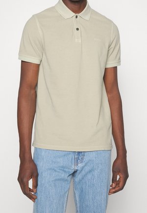 Koszulka polo w jasnym beżowym kolorze z bawełny, z kołnierzykiem, zapięciem na dwa guziki i ribbowymi akcentami na rękawach; małe logo na piersi. Noszona z jasnoniebieskimi dżinsami.