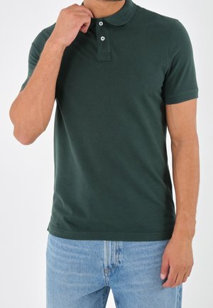 Man die de kraag van een donkergroen polo met korte mouwen verstelt, draagt lichtblauwe spijkerbroek, tegen een effen witte achtergrond.