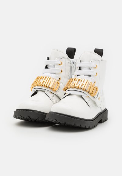 Valged patendi nahast pahkluu saapad kuldse logo rihmaga, ümar varvas, must kummitald ja valged paelad; külgedel on hõbedane tõmblukk.