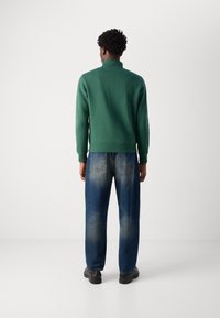 Zelená mikina s vysokým límcem, doplněná světle modrými volnými džínami s vybledlou texturou. Černé boty dokončují outfit.