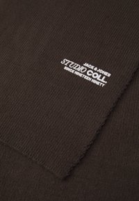 Tejido de punto acanalado marrón que presenta una etiqueta bordada en blanco que dice "JACK & JONES STUDIO COLL DESDE MIL NOVECIENTOS NOVENTA".